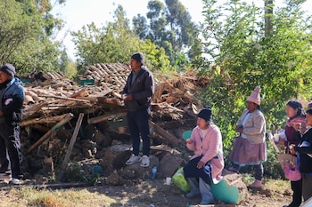 Grupo de hombres, mujeres y niños de pie y sentados junto a escombros de una vivienda, con árboles y vegetación al fondo bajo un cielo claro