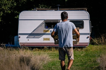 Hombre de espaldas con camiseta gris y pantalón corto, lleva una caja de madera. Al fondo, autocaravana blanca con rayas marrones, árboles y vegetación