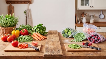 Vegetales frescos enteros y troceados a la izquierda y bolsas con verduras y frutos rojos congelados a la derecha sobre madera.