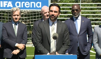 Un hombre barbudo habla en un podio con un micrófono, flanqueado por otros hombres en traje, frente a una portería de fútbol y un cartel de 'FIFA arena'