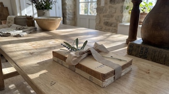 Mesa de madera con dos libros antiguos atados con cinta y decorados con lavanda y hojas de olivo. Al fondo, un cuenco, un tarro, un libro y una ventana.