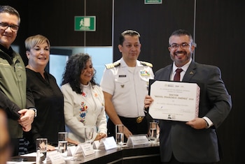 Un hombre con traje sonríe mientras sostiene un diploma, flanqueado por dos mujeres y un oficial del ejército mexicano en uniforme blanco, en un evento