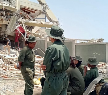 Varias mujeres con uniforme militar verde y sombreros verdes están paradas o sentadas junto a un edificio derrumbado y escombros bajo un cielo claro