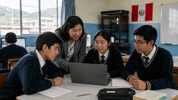 Una docente y tres estudiantes en uniforme interactúan frente a un ordenador portátil en un aula. Hay una bandera de Perú y un estudiante adicional al fondo