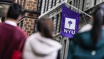 Bandera morada de NYU con logo de antorcha blanca, ondeando en la fachada de un edificio de piedra y ladrillo. Personas borrosas en primer plano
