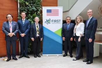 Seis personas con traje formal posan junto a un panel con la bandera de Estados Unidos y el logotipo del programa FIRST