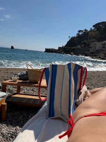 Visão em primeira pessoa de uma pessoa deitada em uma praia de calhau usando um biquíni vermelho, uma bolsa de praia com um toque especial e o mar da Sicília ao fundo.