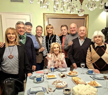 Una docena de personas posan de pie y sentadas alrededor de una mesa puesta con vajilla, copas, postres, tartas y bocadillos