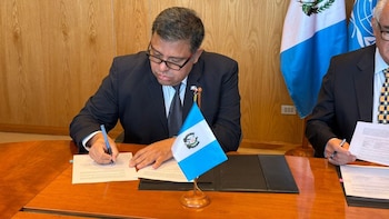 Tres funcionarios, dos hombres y una mujer, sentados en una mesa de madera con banderas de Guatemala y la ONU al fondo, y pequeñas banderas de escritorio