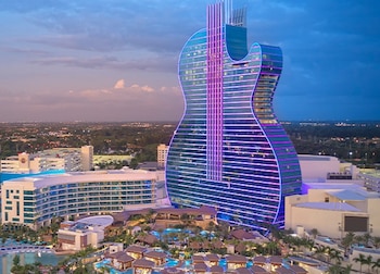 Vista aérea do hotel Seminole Hard Rock em forma de guitarra, iluminado por luzes roxas e azuis, próximo a uma piscina, palmeiras e prédios ao entardecer.