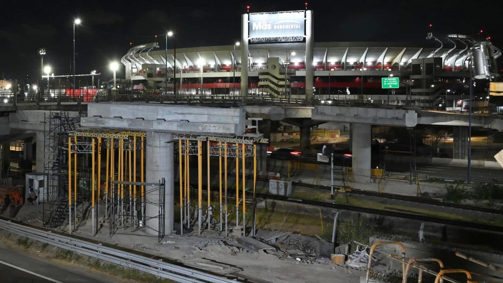 Las obras de construcción del Puente Labruna continúan durante la noche, mostrando su estructura en desarrollo frente al Estadio Monumental de Buenos Aires.