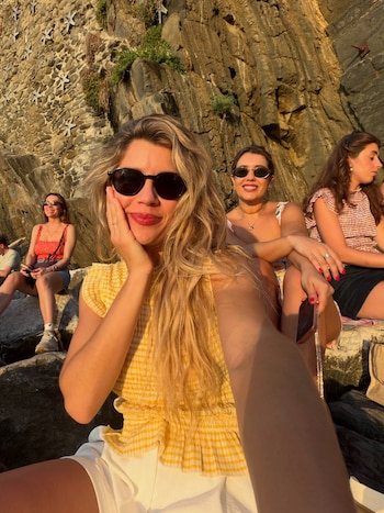 Cuatro mujeres sentadas en rocas junto a un muro de piedra. Una mujer en primer plano con gafas de sol sonríe a cámara. Hay vegetación