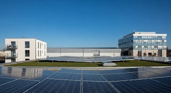 Vista de paneles solares en primer plano bajo cielo azul, con un edificio residencial blanco, una nave industrial gris y un edificio de oficinas al fondo.