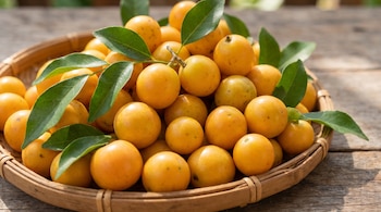 Una cesta de mimbre contiene varios frutos redondos de color amarillo anaranjado, acompañados de hojas verdes, sobre una mesa de madera.