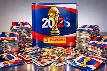 Álbum Panini de la Copa Mundial FIFA 2026 con logo oficial y Panini en portada, rodeado de pilas de sobres de figuritas y cromos dispersos en una superficie brillante.