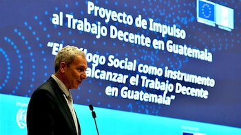 Hombre en traje habla por micrófono. Detrás, pantalla azul con el título del 'Proyecto de Impulso al Trabajo Decente en Guatemala' y dos banderas