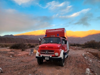 Camión rojo Mercedes-Benz con balizas azules y estructura de motorhome, estacionado en terreno árido. Fondo de montañas iluminadas por el sol y cielo azul con nubes
