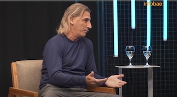 Hombre de cabello largo y camisa azul, sentado en una silla, gesticulando. A su derecha, dos copas de agua sobre una mesa, con fondo oscuro y luces de neón azules
