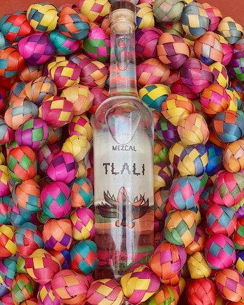 Una botella de vidrio transparente de mezcal Tlali con corcho. Rodeada por una gran cantidad de esferas pequeñas, tejidas, de varios colores