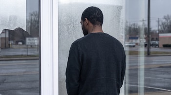 Un hombre de espaldas con suéter oscuro mira por una ventana que combina un panel de vidrio texturizado con otro transparente hacia el exterior.