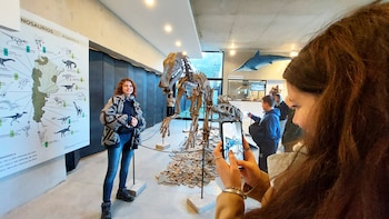 Una mujer sonriente junto a un mapa de dinosaurios de Argentina. Otra persona la fotografía con un celular. Detrás, un esqueleto de dinosaurio y un pez colgando