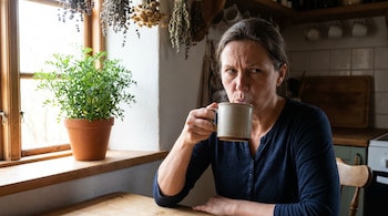 Una mujer de mediana edad con cabello oscuro bebe de una taza beige con una expresión fruncida. Detrás, una planta verde en maceta en el alféizar de una ventana.