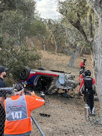 Un auto de carreras volcado entre árboles detrás de una cerca metálica, junto a personal de emergencia y un comisario de pista