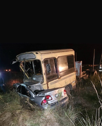 Un automóvil plateado destrozado está empotrado en la parte trasera de un autobús o camión amarillo oxidado en una zanja con pasto alto, de noche