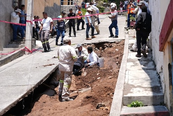 Vista de una calle con una gran zanja de tierra en el centro, donde varios trabajadores realizan reparaciones mientras vecinos observan. Hay cinta de seguridad roja