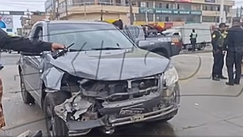 Un automóvil gris con la parte delantera destruida en una vía urbana, con agentes de policía al fondo