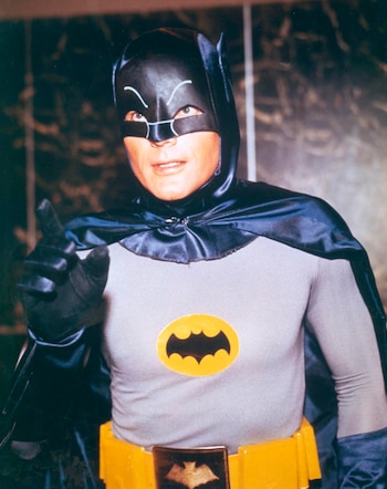 Adam West como Batman con traje gris, capa y máscara negras, cinturón amarillo y logo del murciélago en el pecho, apuntando con la mano derecha