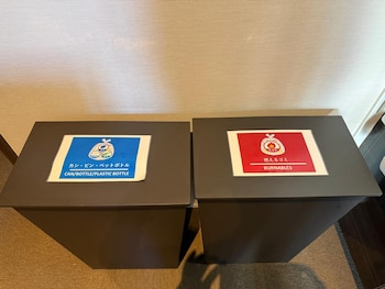 Dos contenedores de basura cuadrados de color gris oscuro con etiquetas. El de la izquierda es azul para reciclaje, el de la derecha es rojo para incinerables