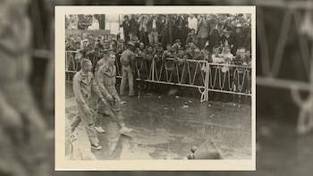 Dos astronautas con trajes de vuelo caminan sobre una superficie mojada, con una valla blanca a su derecha y una multitud de personas tomando fotos detrás