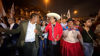 Roberto Sánchez con chaqueta roja y sombrero blanco, junto a un grupo de personas, banderas y edificios iluminados de noche