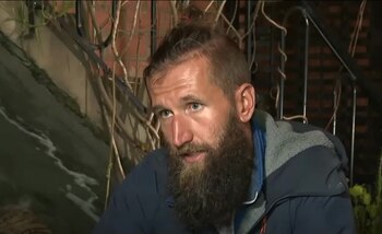 Primer plano de un hombre con barba y cabello recogido. Viste una chaqueta azul. El fondo incluye una pared de ladrillo y ramas de plantas