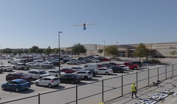 Un dron de ala fija y rotor múltiple en vuelo sobre un estacionamiento abarrotado con cientos de coches y SUV, con un edificio de Walmart en el fondo