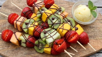 Brochetas de madera con trozos de fresa, kiwi y melón bañados en chocolate, junto a un cuenco con helado de limón y menta sobre una tabla.