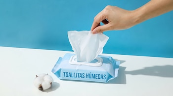 Una mano saca una toallita blanca de un paquete celeste con el texto TOALLITAS HÚMEDAS junto a un capullo de algodón.