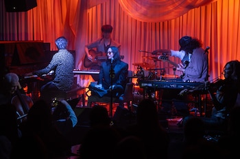 Integrantes de una banda musical tocan piano, guitarra, batería y violín en un escenario con cortinas rojas e iluminación cálida