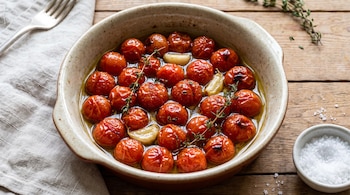 Recipiente de cerámica con tomates cherry, aceite, ajos y tomillo, junto a un tenedor, un paño de tela y un cuenco con sal sobre madera.