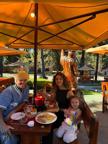 Alex Caniggia, Melody Luz y su hija Venezia sentados en una mesa de madera al aire libre bajo un parasol naranja. Hay un fondue y comida