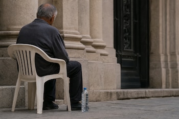 Un hombre de espaldas sentado en una silla blanca de plástico con una botella de agua en el suelo frente a un edificio de piedra.