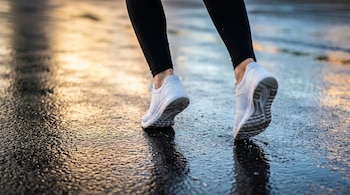 Piernas con pantalón deportivo negro y zapatillas blancas avanzan sobre asfalto urbano mojado con reflejos dorados y azules del amanecer.