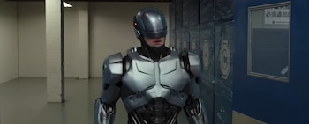 Primer plano de Robocop de pie, con traje robótico gris y negro, en un almacén con paredes blancas, cajas apiladas y una ventana lateral