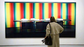 Espalda de una persona con gabardina y bolso frente a una gran obra de arte con fondo de franjas de colores verticales y una mesa con sillas