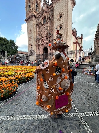 Vista trasera de persona con traje marrón decorado con elementos de Día de Muertos y sombrero con calaveras, frente a iglesia de cantera rosa y campo de cempasúchil