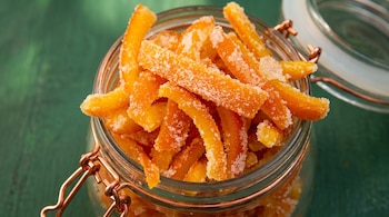 Tiras de cáscara de naranja confitada con azúcar dentro de un frasco de vidrio con cierre metálico, sobre un fondo verde.