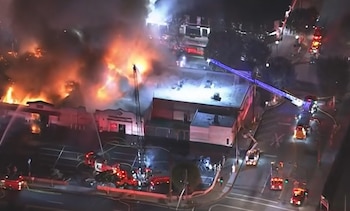 Vista aérea nocturna de un edificio envuelto en llamas y humo, rodeado por camiones de bomberos con escaleras y mangueras