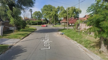 Una calle de asfalto con la inscripción Los Cipreses, bordeada por árboles, césped, casas y un automóvil rojo al fondo