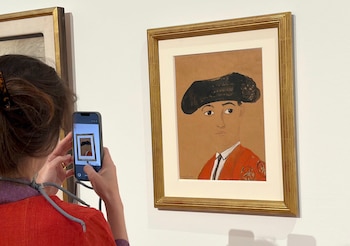 Una persona de espaldas con blusa roja sostiene un teléfono móvil, fotografiando un cuadro con marco dorado. El cuadro muestra el retrato de un hombre